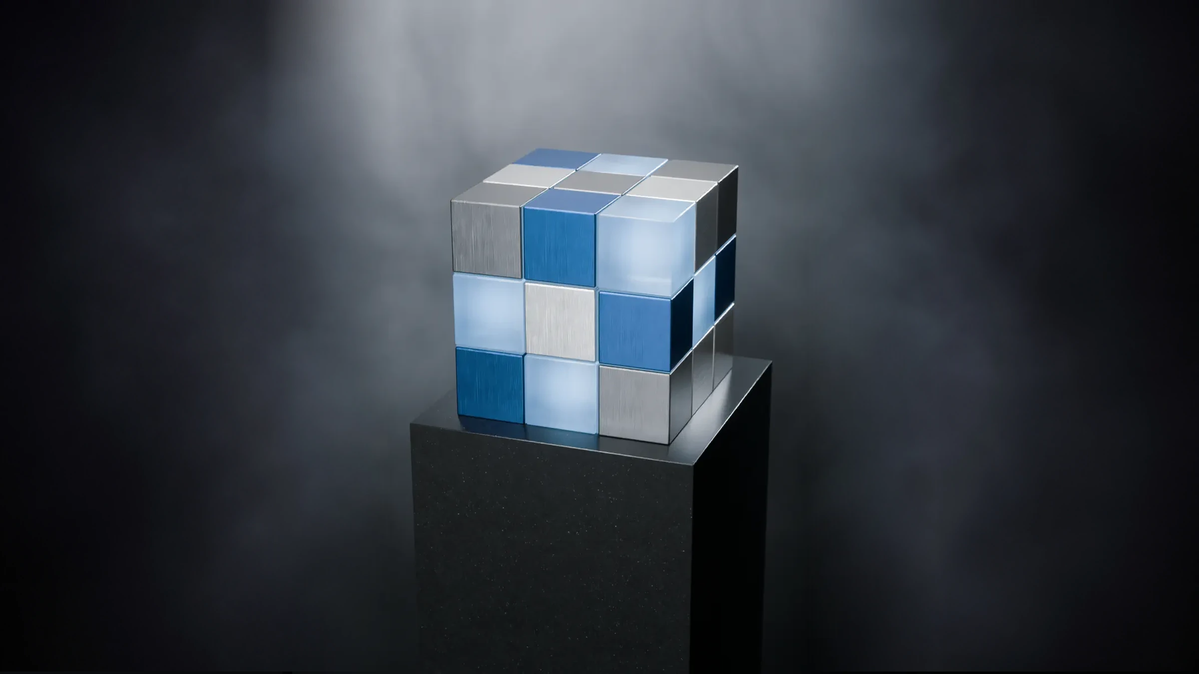A solved cube of brushed silver, navy and frosted glass on a stone plinth