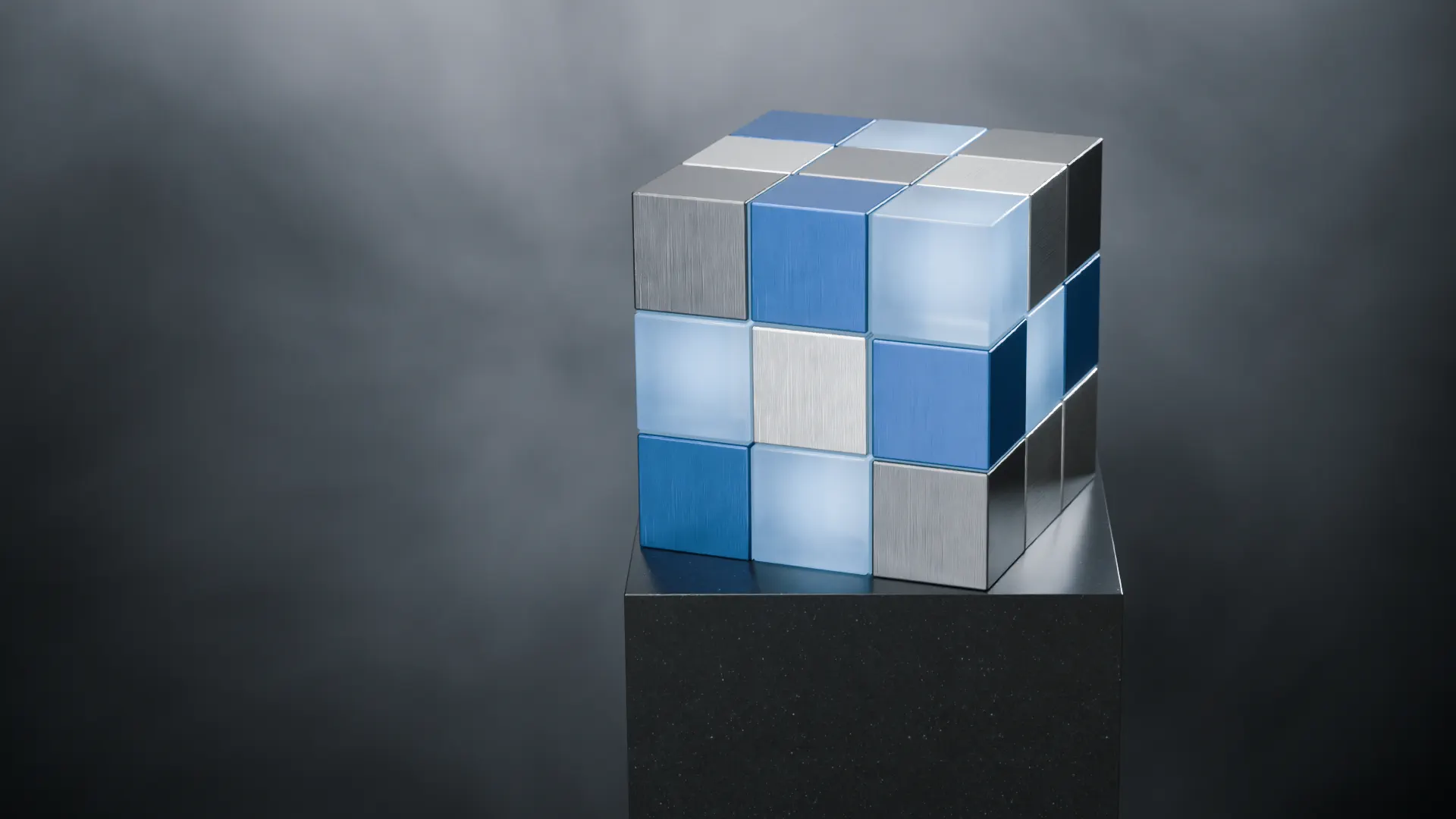 The solved BTI cube in brushed aluminium, blue anodised metal and frosted glass, at rest on a black stone plinth.